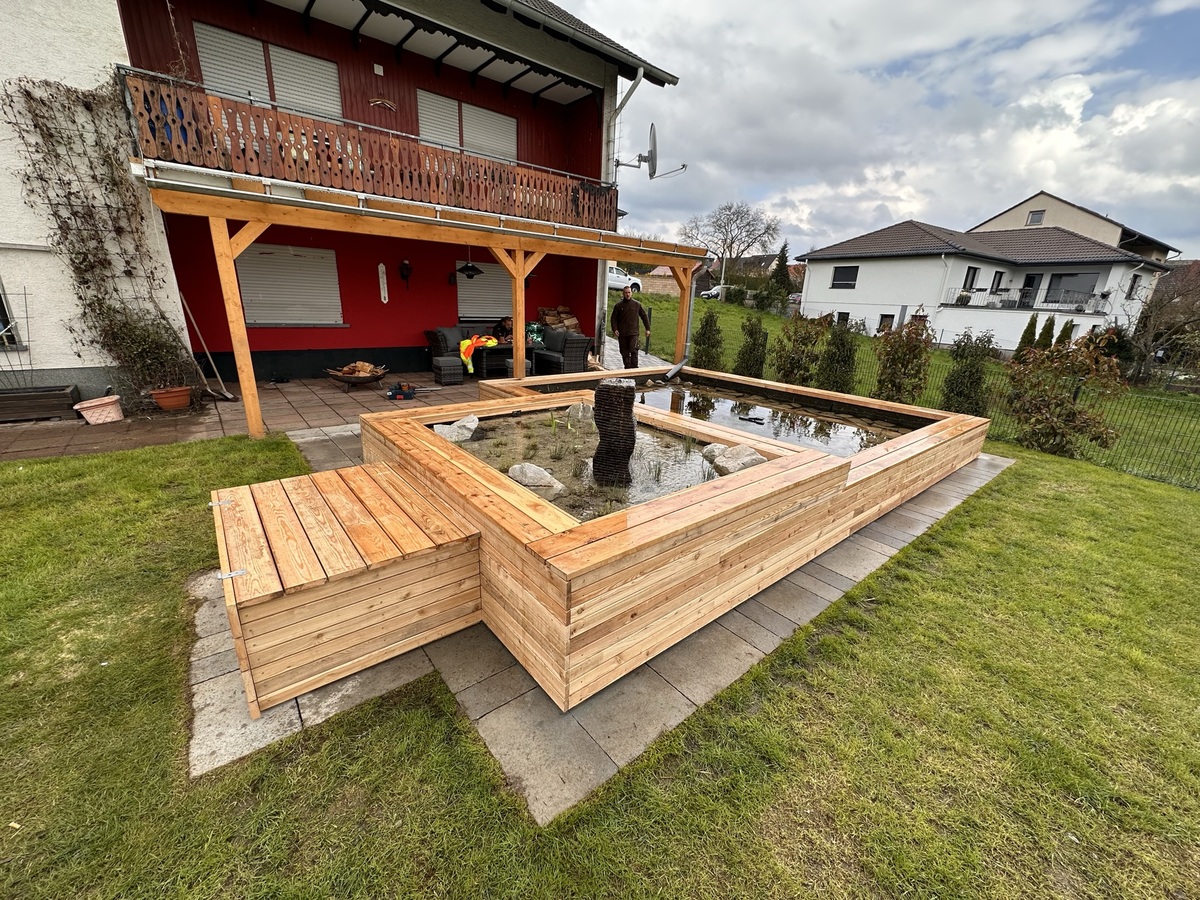 Holzteichbau im Garten vor einem Haus mit überdachter Terrasse und Wolkenhimmel
