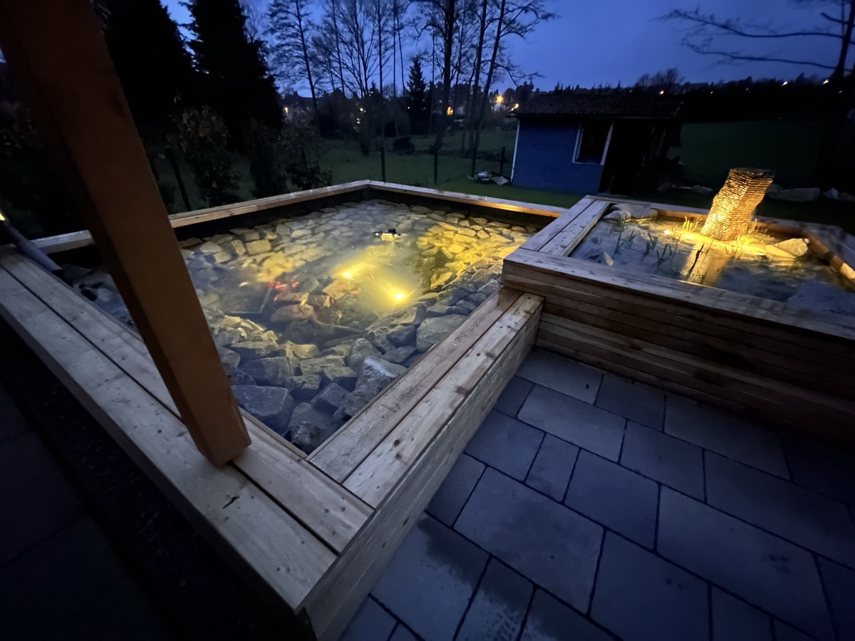 Ein beleuchteter Gartenbrunnen aus Holzrahmen und Steinen in der Dämmerung.