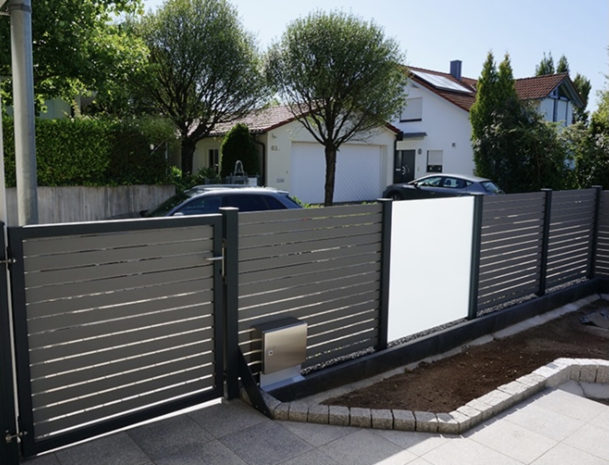 Ein grauer Zaun mit modernem Design steht vor einem Haus mit zwei Bäumen.