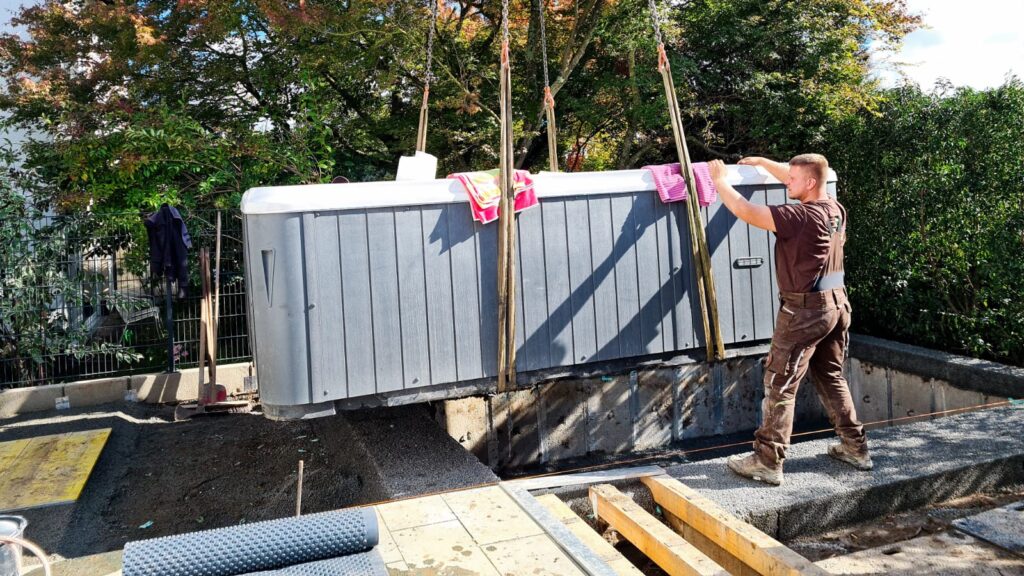 Eine Person hebt konzentriert einen großen grauen Kasten in einen Garten.