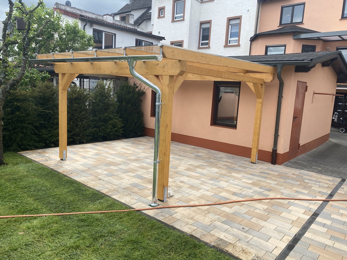 Ein hölzerner Carport steht auf gepflastertem Boden vor einem Haus mit beigem Putz und brauner Umrandung.