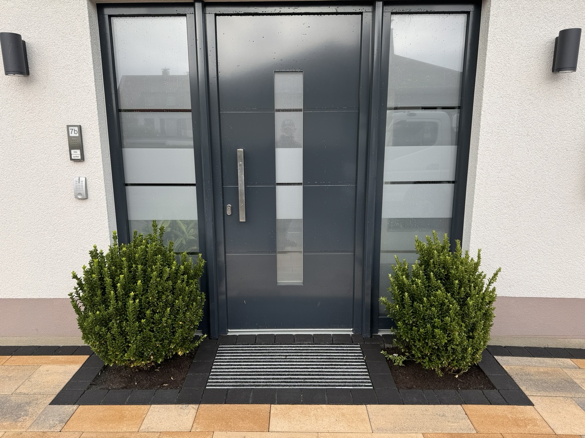 Eine moderne, graue Eingangstür flankiert von zwei grünen Sträuchern.