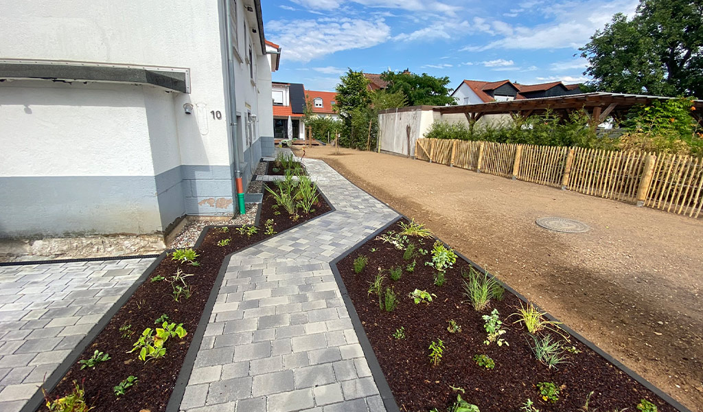 Ein neu angelegter Gartenweg aus grauen Pflastersteinen führt an bunten Beeten entlang.