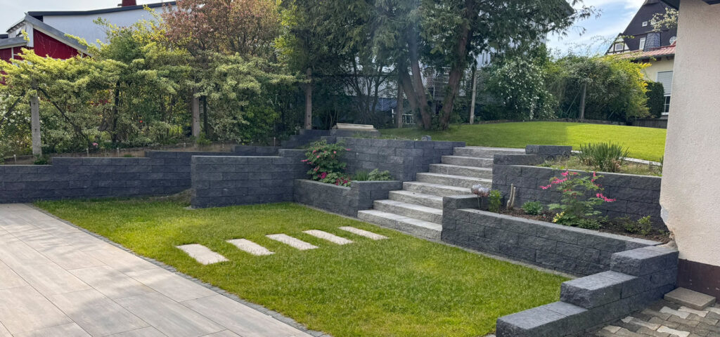 Ein gepflegter Garten mit modernen grauen Steinmauern und Stufen in der Sonne.